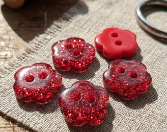 May include: Four flower-shaped buttons with a red glitter finish and one solid red flower-shaped button, each featuring two central holes. These five buttons are arranged on a piece of light beige fabric near a spool of red thread and scissors.