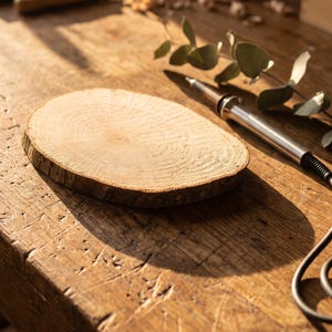 May include: A circular, light-coloured wooden slice with visible grain patterns and rough bark edges, positioned next to a metallic wood-burning tool with a black cord and a small green leafy branch on a weathered, textured wooden surface.