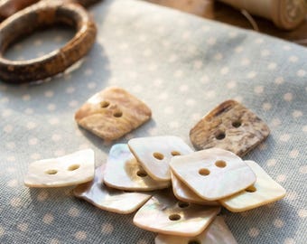 May include: A collection of square, two-hole buttons with a pearlescent, off-white finish and some with brown marbled patterns. The buttons are scattered on a light blue fabric featuring a white polka dot pattern, with sewing tools nearby.