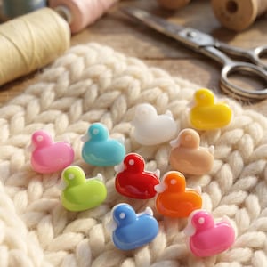 May include: A collection of small, glossy, duck-shaped buttons in various colours including pink, light blue, white, yellow, beige, red, lime green, and orange, arranged on a thick, cream-coloured knitted fabric with sewing supplies in the background.