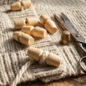 May include: A collection of light-coloured wooden toggle buttons arranged on a beige knitted fabric. Beside the buttons are a pair of vintage metal scissors and a small brass thimble. The buttons feature a smooth, cylindrical shape with a centre groove.
