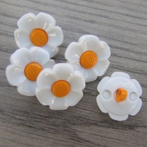 May include: Five white plastic buttons shaped like daisies with orange centres. One button has a shank on the back.