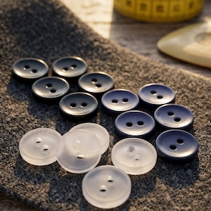 May include: A collection of round, two-hole buttons in solid black and translucent white, arranged on a textured gray fabric surface. The buttons are circular with a smooth finish, featuring two centered holes for attachment to garments or crafts.