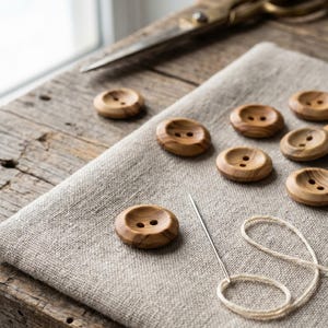 May include: Wooden buttons with two holes, displayed on a beige linen cloth. A needle with white thread is placed near the buttons. Vintage scissors are in the background, indicating a sewing or craft project.