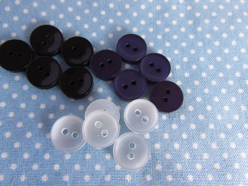 May include: A collection of round, two-hole buttons in three distinct colors: solid black, deep purple, and translucent white. The buttons are arranged in a loose cluster, showcasing their smooth, circular surfaces and the two centered holes.