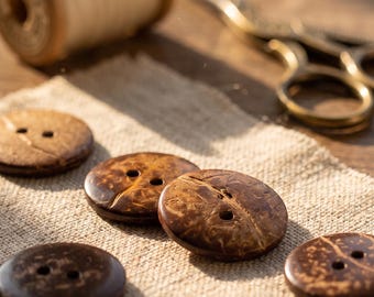 May include: Five round, dark brown buttons with visible natural wood grain patterns and two holes in the center. The buttons are arranged on a piece of light tan fabric, accompanied by a wooden spool of white thread and metal sewing scissors.