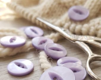May include: A collection of round, light purple buttons with a central eye-shaped indentation and two holes. The buttons are scattered alongside a pair of silver metal scissors and a ball of thick, cream-colored yarn on a light wooden surface.