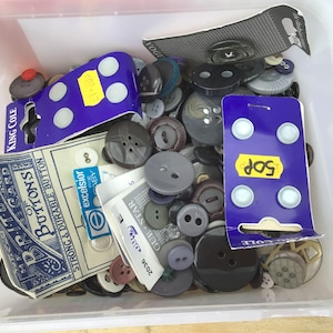 Peut inclure: Un récipient en plastique transparent rempli de boutons de différentes couleurs, allant du gris au marron et au noir. Certains boutons sont encore attachés à leur emballage d'origine, avec des étiquettes "BRITON BUTTONS".