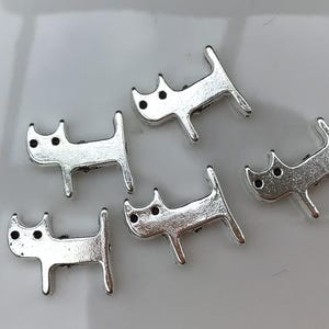 May include: Five silver charm pendants shaped like cats. Each charm has a simple design with two small circles for eyes.