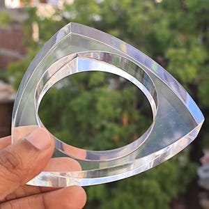 Op de afbeelding: Transparante acryl armband met een driehoekige buitenkant en een cirkelvormige binnenopening. Het heldere materiaal reflecteert licht. De armband wordt in een hand gehouden tegen een vage groene achtergrond.