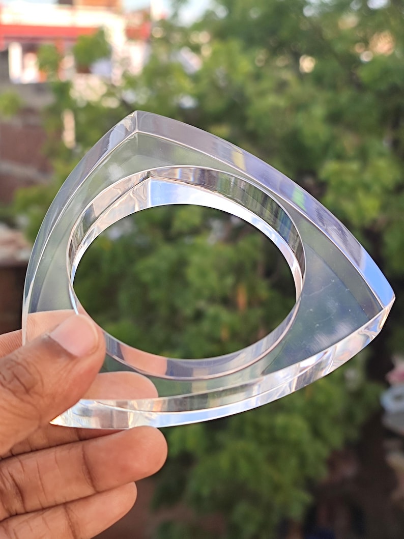 Op de afbeelding: Transparante acryl armband met een driehoekige buitenkant en een cirkelvormige binnenopening. Het heldere materiaal reflecteert licht. De armband wordt in een hand gehouden tegen een vage groene achtergrond.
