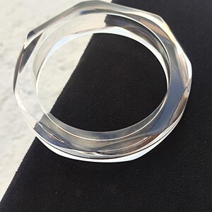 Puede incluir: Brazalete transparente y geom&eacute;trico. El brazalete transparente tiene un dise&ntilde;o hexagonal y est&aacute; hecho de un material transparente y brillante. El brazalete se muestra sobre una superficie negra.