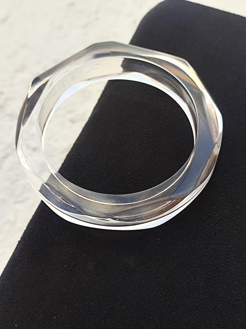 Puede incluir: Brazalete transparente y geom&eacute;trico. El brazalete transparente tiene un dise&ntilde;o hexagonal y est&aacute; hecho de un material transparente y brillante. El brazalete se muestra sobre una superficie negra.