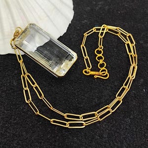 Puede incluir: Un collar dorado con un colgante rectangular de cristal transparente. El colgante está engastado en un marco dorado. La cadena está hecha de eslabones alargados dorados. Fondo negro con una concha blanca.