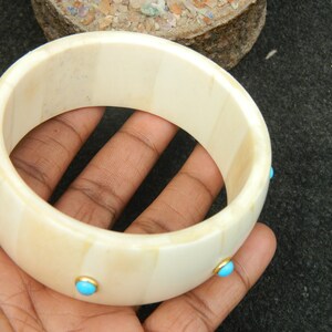 May include: A white bone bangle bracelet with turquoise stones set in gold. The bracelet is about 2 inches wide and has a smooth, polished finish.