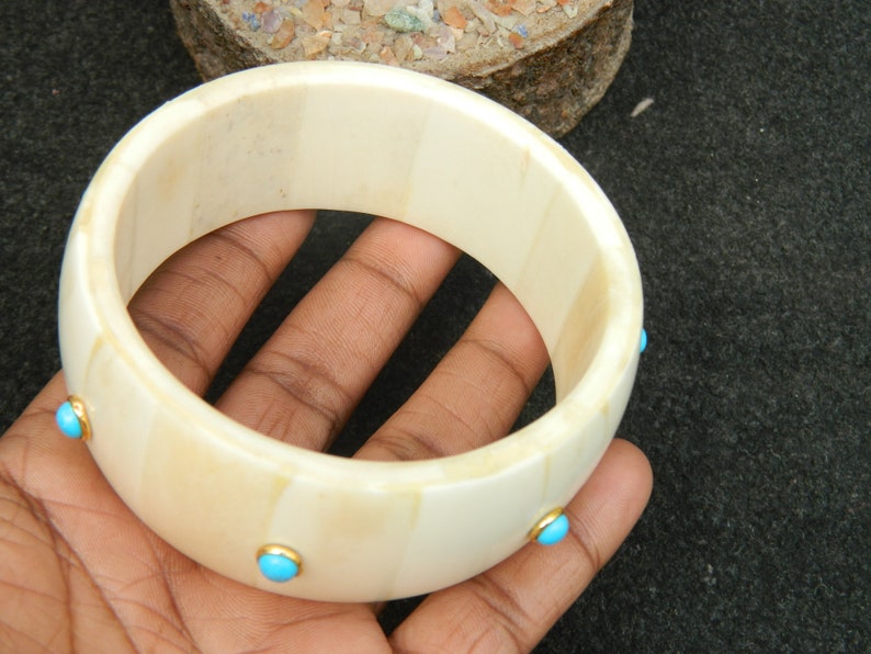May include: A white bone bangle bracelet with turquoise stones set in gold. The bracelet is about 2 inches wide and has a smooth, polished finish.