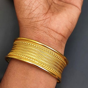 May include: A gold bangle bracelet with a textured design. The bracelet is made of multiple bands with a beaded edge.
