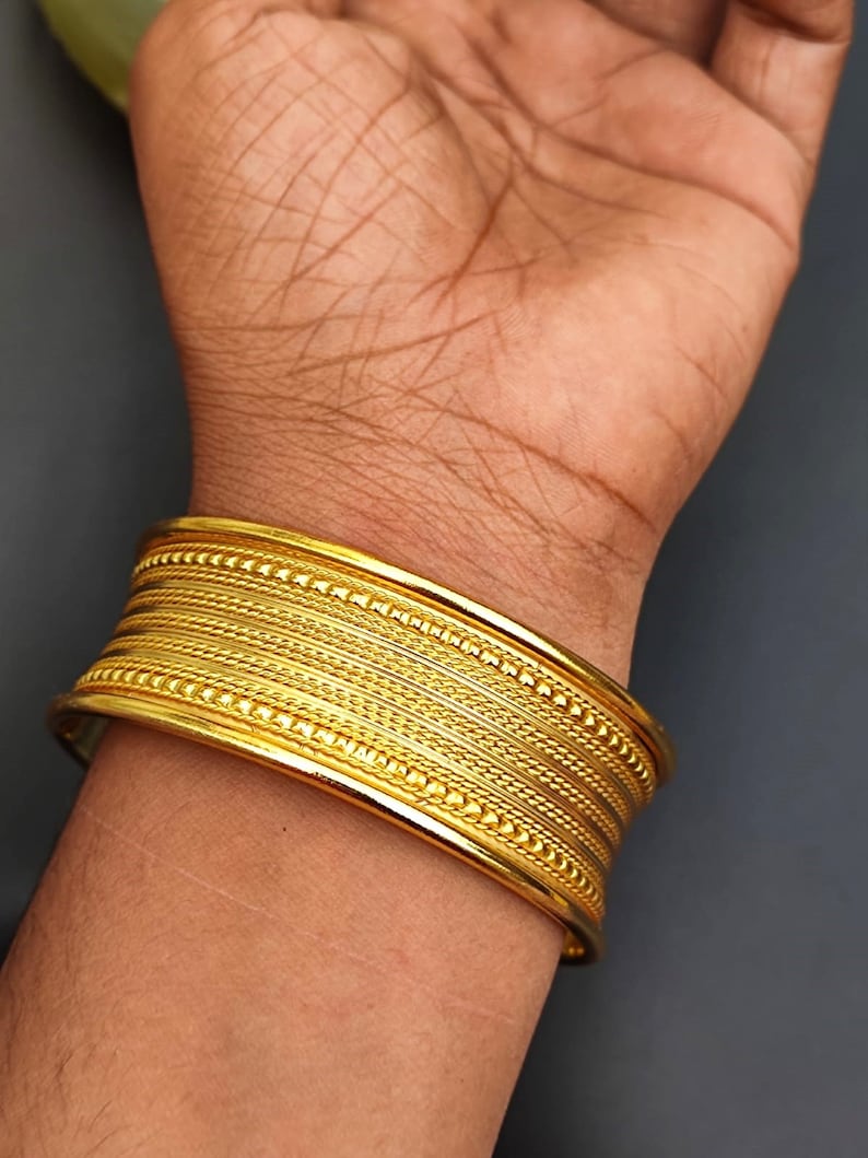 May include: A gold bangle bracelet with a textured design. The bracelet is made of multiple bands with a beaded edge.