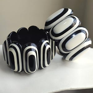 May include: Two black and white striped bracelets. One is a wide, scalloped cuff, and the other is a series of oval links. The bracelets are made of a glossy material and are set against a neutral background.