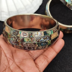 Puede incluir: Un brazalete de plata con un mosaico de piezas de concha de abulón iridiscente. Las piezas de concha están dispuestas en un patrón geométrico y tienen una variedad de colores, incluyendo verde, azul y dorado.