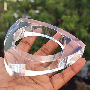 Op de afbeelding: Transparante acryl armband met een driehoekig ontwerp. De armband heeft een geometrische vorm met afgeronde randen en een cirkelvormige opening in het midden. Het transparante materiaal laat licht door, waardoor een moderne en minimalistische uitstraling ontstaat.