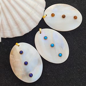 May include: Three oval-shaped pendants with a mother-of-pearl finish, each adorned with three small gemstones. The gemstones are amber, turquoise, and deep blue. Each pendant has a gold-colored bail for hanging.