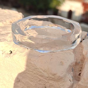 Puede incluir: Un brazalete de vidrio transparente y facetado. El brazalete tiene un dise&ntilde;o geom&eacute;trico con m&uacute;ltiples lados, que refleja la luz. El material transparente permite ver a trav&eacute;s del centro. El brazalete est&aacute; sobre una superficie texturizada de color claro.