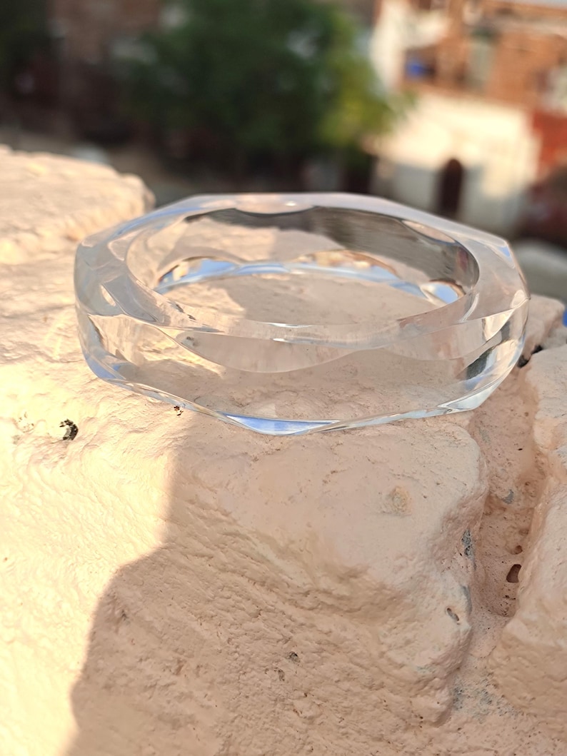 Puede incluir: Un brazalete de vidrio transparente y facetado. El brazalete tiene un dise&ntilde;o geom&eacute;trico con m&uacute;ltiples lados, que refleja la luz. El material transparente permite ver a trav&eacute;s del centro. El brazalete est&aacute; sobre una superficie texturizada de color claro.