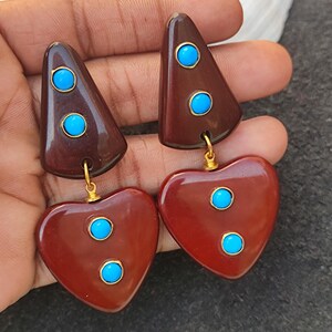 May include: A pair of brown and red heart-shaped earrings with turquoise stones. The earrings are made of resin and have a gold-tone finish.