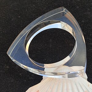 Op de afbeelding: Een heldere, driehoekige acrylarmband. De armband ligt op een witte zeeschelp tegen een donkere achtergrond. De armband heeft een geometrisch ontwerp en een gepolijste afwerking, die het licht reflecteert. Binnendiameter ongeveer 6 cm.