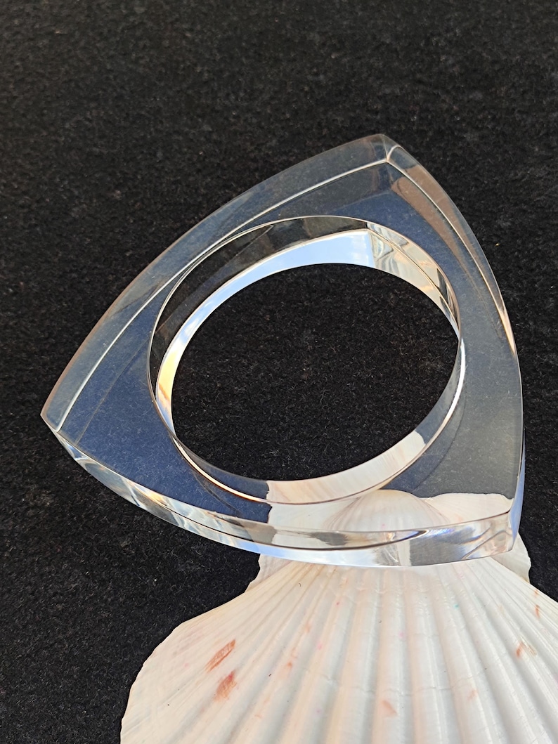 Op de afbeelding: Een heldere, driehoekige acrylarmband. De armband ligt op een witte zeeschelp tegen een donkere achtergrond. De armband heeft een geometrisch ontwerp en een gepolijste afwerking, die het licht reflecteert. Binnendiameter ongeveer 6 cm.