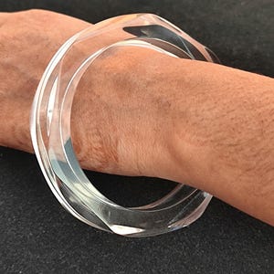 Puede incluir: Brazalete de acr&iacute;lico transparente. El brazalete tiene un dise&ntilde;o geom&eacute;trico transparente con forma cuadrada redondeada. El brazalete se lleva en la mu&ntilde;eca y tiene un acabado liso y pulido. La joya es un accesorio moderno.