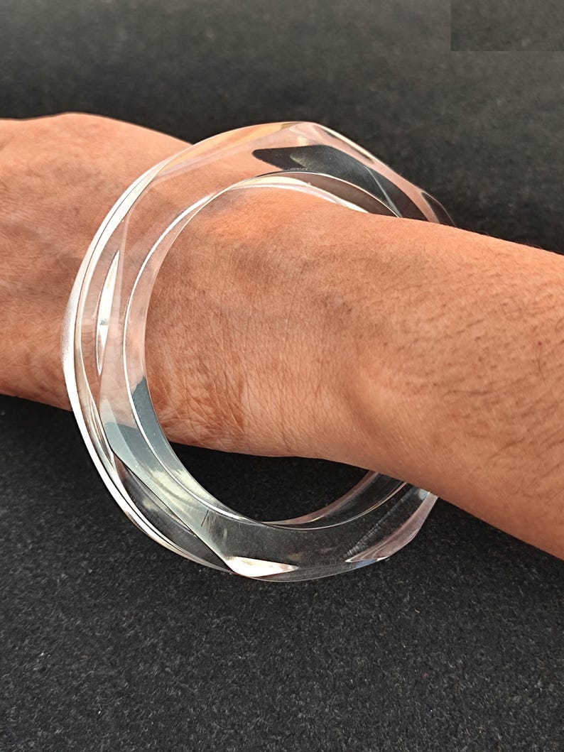 Puede incluir: Brazalete de acr&iacute;lico transparente. El brazalete tiene un dise&ntilde;o geom&eacute;trico transparente con forma cuadrada redondeada. El brazalete se lleva en la mu&ntilde;eca y tiene un acabado liso y pulido. La joya es un accesorio moderno.