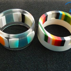 May include: Two colorful resin bangle bracelets. The first bracelet is clear with orange, white, blue, and black geometric shapes. The second bracelet is white with a multi-colored stripe pattern.