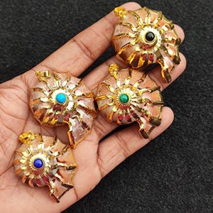 May include: Four gold-coloured nautilus shell pendants, each with a different gemstone centre: blue, green, black, and turquoise. The pendants have an intricate, textured design, with a detailed finish.