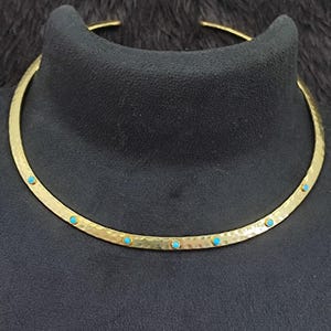 Könnte beinhalten: Ein goldfarbenes Choker-Halsband mit gehämmerten Textur und eingelegten türkisfarbenen Steinen. Die Halskette wird auf einem schwarzen Mannequinkopf präsentiert, um ihr Design hervorzuheben. Das Schmuckstück hat ein schlichtes, elegantes Design.