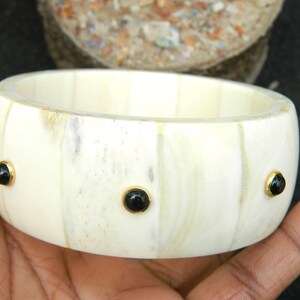 May include: A white bone bangle bracelet with black gemstone accents. The bracelet is made of natural bone and has a polished finish.
