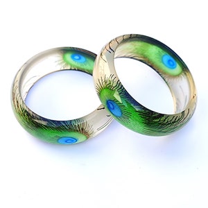 May include: Two clear resin bangles with peacock feather designs. The bangles are translucent and feature green and blue peacock feathers with a white background.