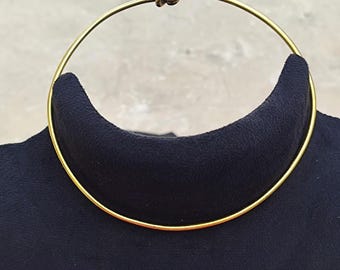 May include: A gold-colored choker necklace. The necklace is a simple, elegant design, featuring a thin, circular band that sits close to the neck. A small, decorative element is at the front of the necklace.
