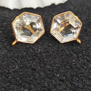 May include: A pair of gold-toned stud earrings. Each earring features a clear, faceted, hexagonal crystal set within a gold frame. The earrings are displayed on a dark gray textured surface, with a white seashell in the background.