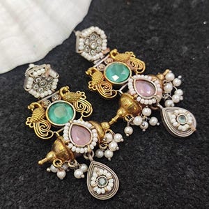 Può includere: Orecchini dorati ornati con pietre preziose verdi e rosa, impreziositi da piccole perle bianche. Gli orecchini presentano un design vintage dettagliato con un pendente a forma di goccia. È presente anche un orecchino a lobo abbinato.