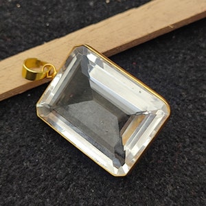 May include: A clear quartz crystal pendant with a gold-toned frame and a bail. The pendant is rectangular in shape and has a faceted design.