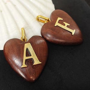 May include: Two heart-shaped pendants with gold-colored letters "A" and "F" on a dark brown wood surface. Each pendant has a gold-colored bail. The background includes a white seashell and a black surface.