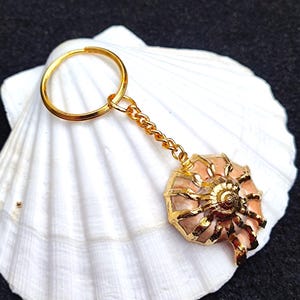 May include: A gold-tone keychain with a pink seashell charm. The charm has a detailed design and a gold-tone finish.