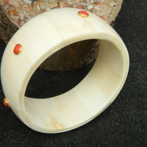 May include: A white bone bangle bracelet with three red gemstone accents. The bracelet is made of polished bone and has a smooth, rounded surface.