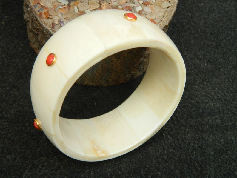 May include: A white bone bangle bracelet with three red gemstone accents. The bracelet is made of polished bone and has a smooth, rounded surface.