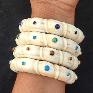 May include: Five ivory bangles with gold accents and colourful gemstones. The bangles are stacked on a person's wrist.
