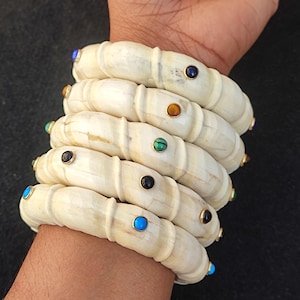 May include: A set of six ivory bangles with colourful gemstone accents. The bangles are stacked on a person's wrist and feature a variety of colours, including blue, green, and brown.