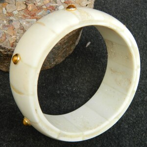 May include: White bone bangle bracelet with gold accents. The bracelet is made of bone and has a smooth, polished finish. The gold accents are small and round, and they are evenly spaced around the bracelet.