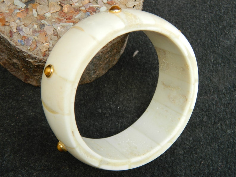 May include: White bone bangle bracelet with gold accents. The bracelet is made of bone and has a smooth, polished finish. The gold accents are small and round, and they are evenly spaced around the bracelet.